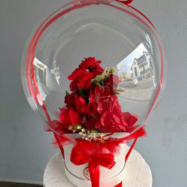 Aranjament floral cu flori roșii în balon transparent, decorat cu panglică roșie, perfect pentru evenimente speciale sau cadouri romantice.