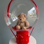Ursuțel de pluș în balon transparent, decor pentru evenimente speciale, cu fundiță roșie, pe suport roșu, ideal pentru aniversări și petreceri tematice.